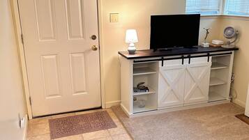 A room with a white door, a television on a wooden stand, a white cabinet with shelves, and a carpeted floor.