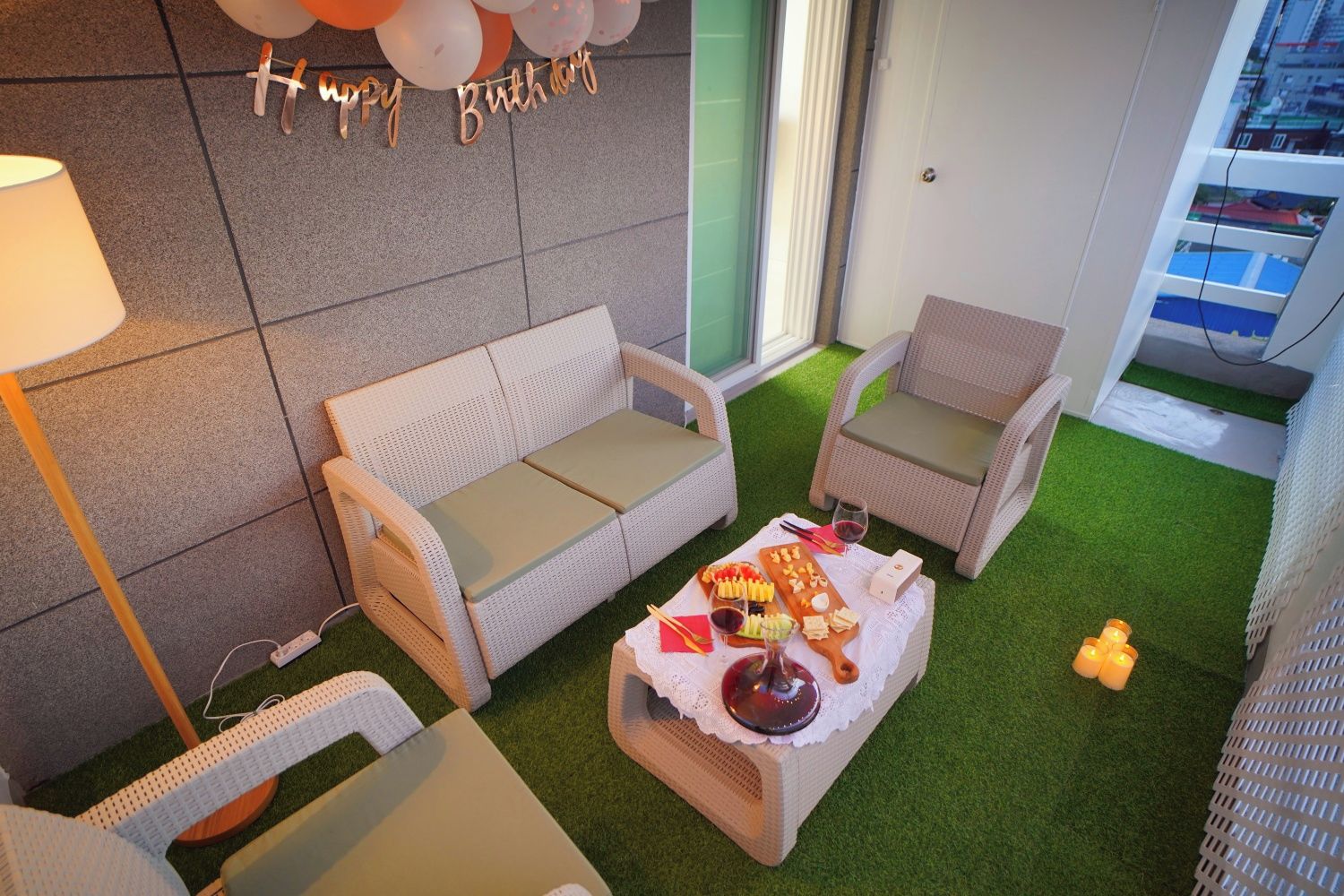 Un rincón al aire libre acogedor con un sofá y sillones, una mesa pequeña con aperitivos y bebidas, y una decoración con un globo de 'Happy Birthday'.
