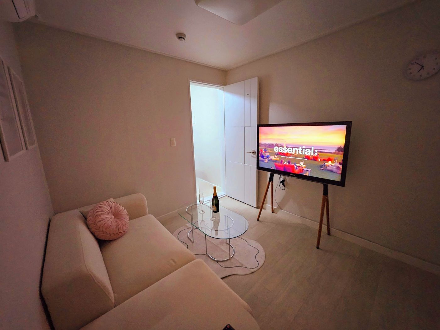 Una sala de estar con un sofá, una mesa de centro de vidrio, un televisor de pantalla plana sobre un mueble bajo y una puerta que da a otra habitación.
