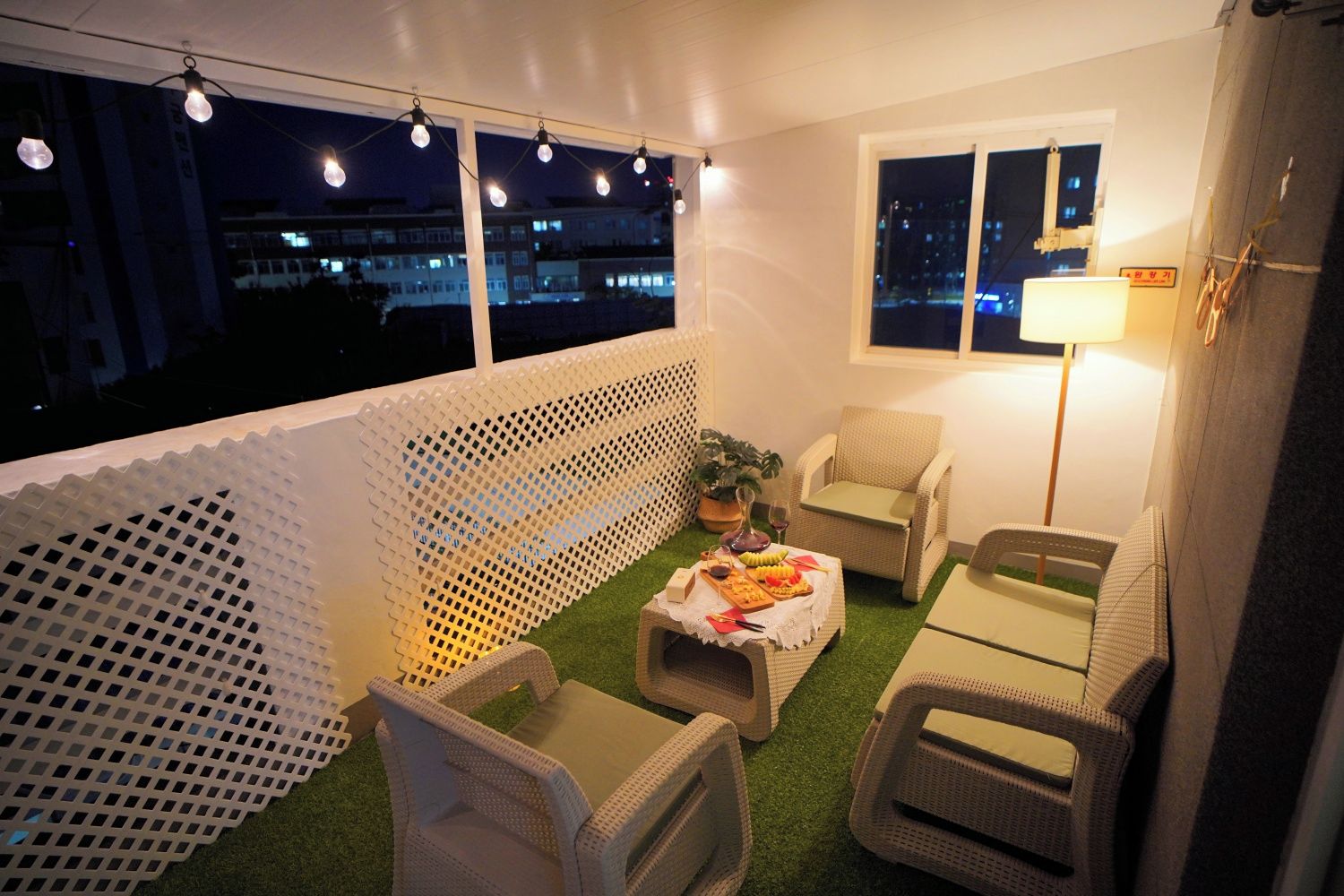 Un balcón con mobiliario exterior, una mesa con comida y bebidas, y vista a la ciudad de noche.