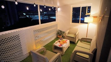 Un balcón con mobiliario exterior, una mesa con comida y bebidas, y vista a la ciudad de noche.