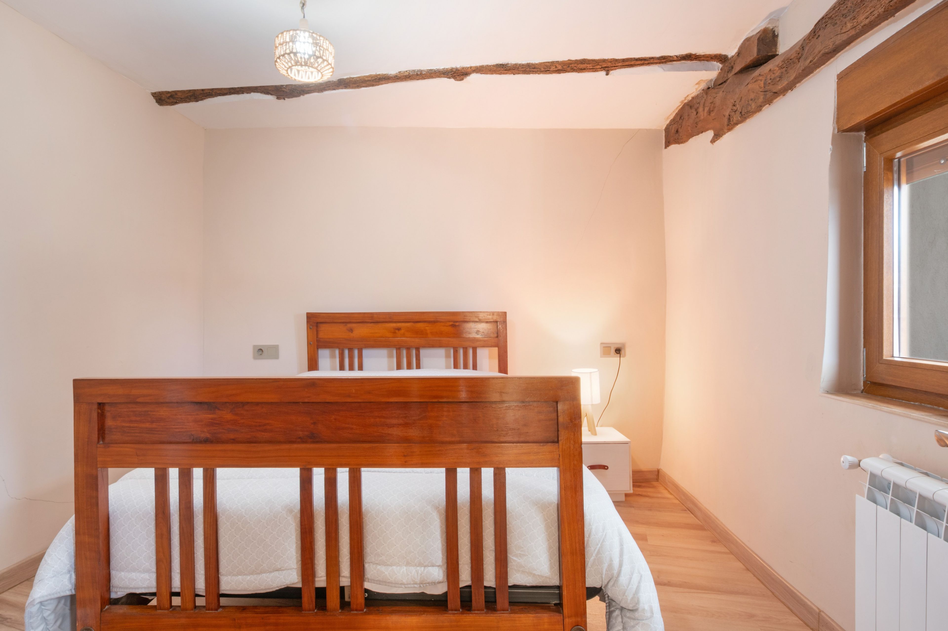 Un dormitorio con una cama de madera, una mesita de noche con lámpara, un radiador y una ventana.