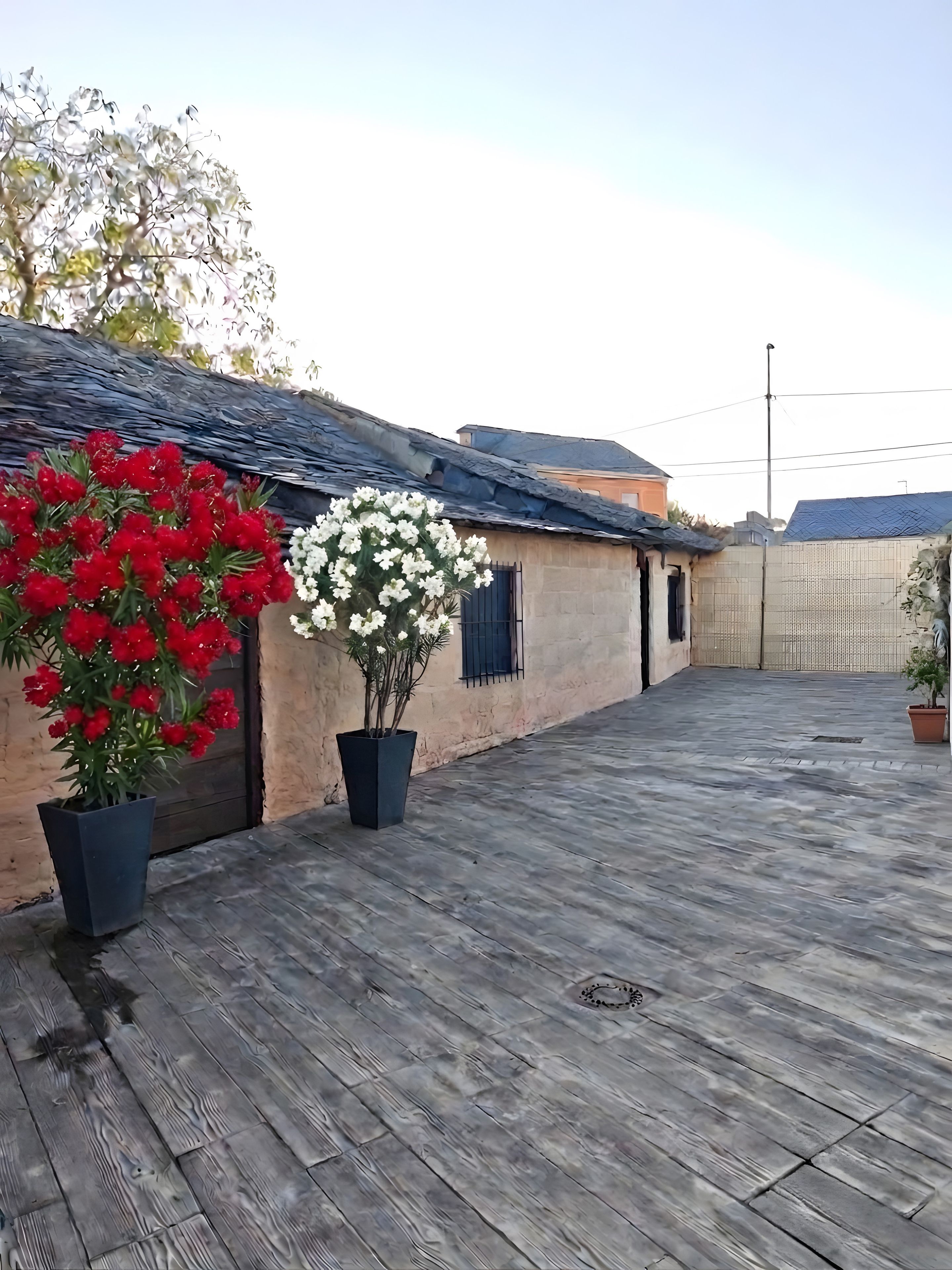 Een binnenplaats met een houten vloer, twee potplanten met rode en witte bloemen, en een gebouw met een pannendak.