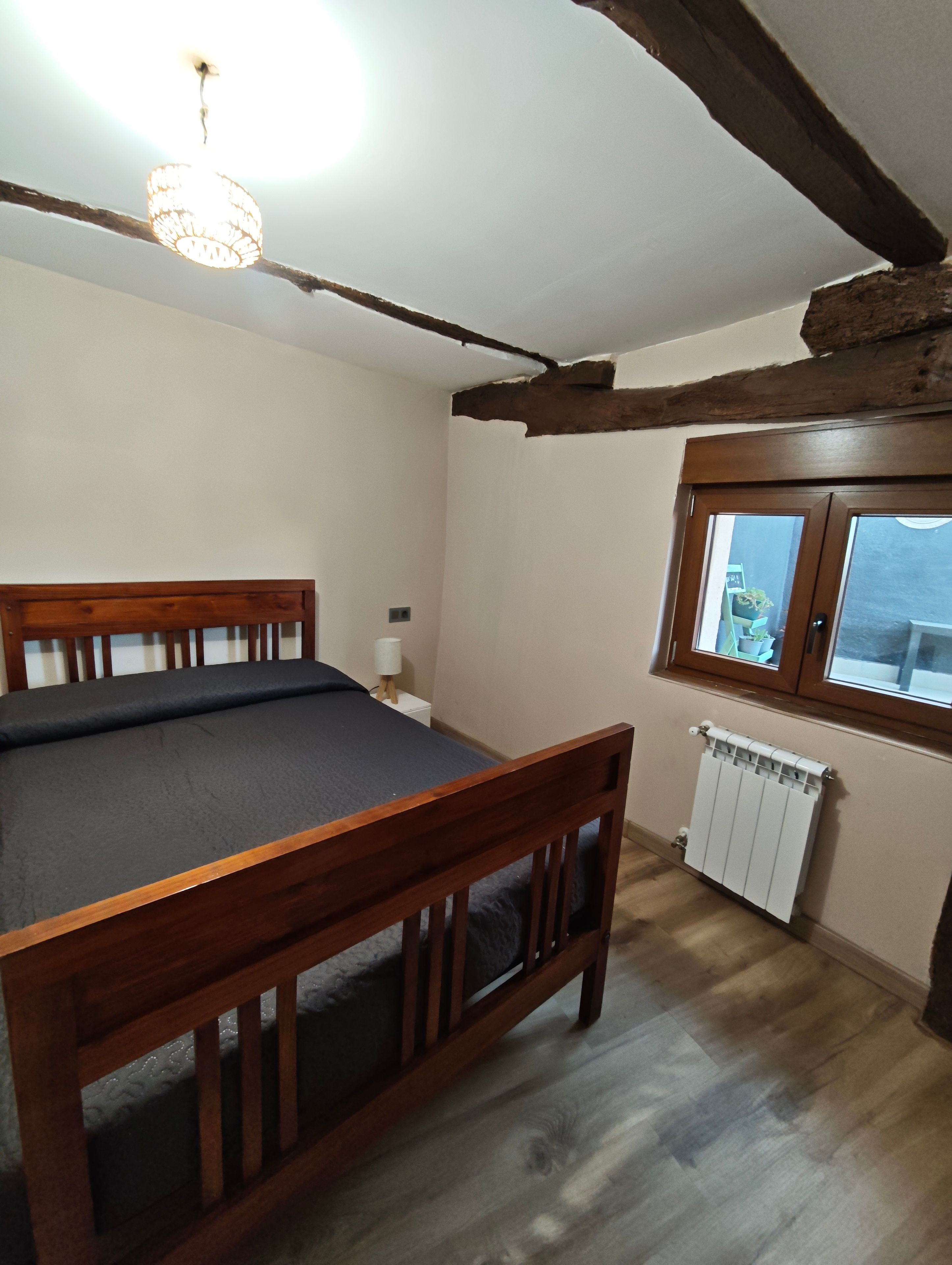 Una habitación con cama de madera, un radiador, una ventana y una lámpara en el techo.