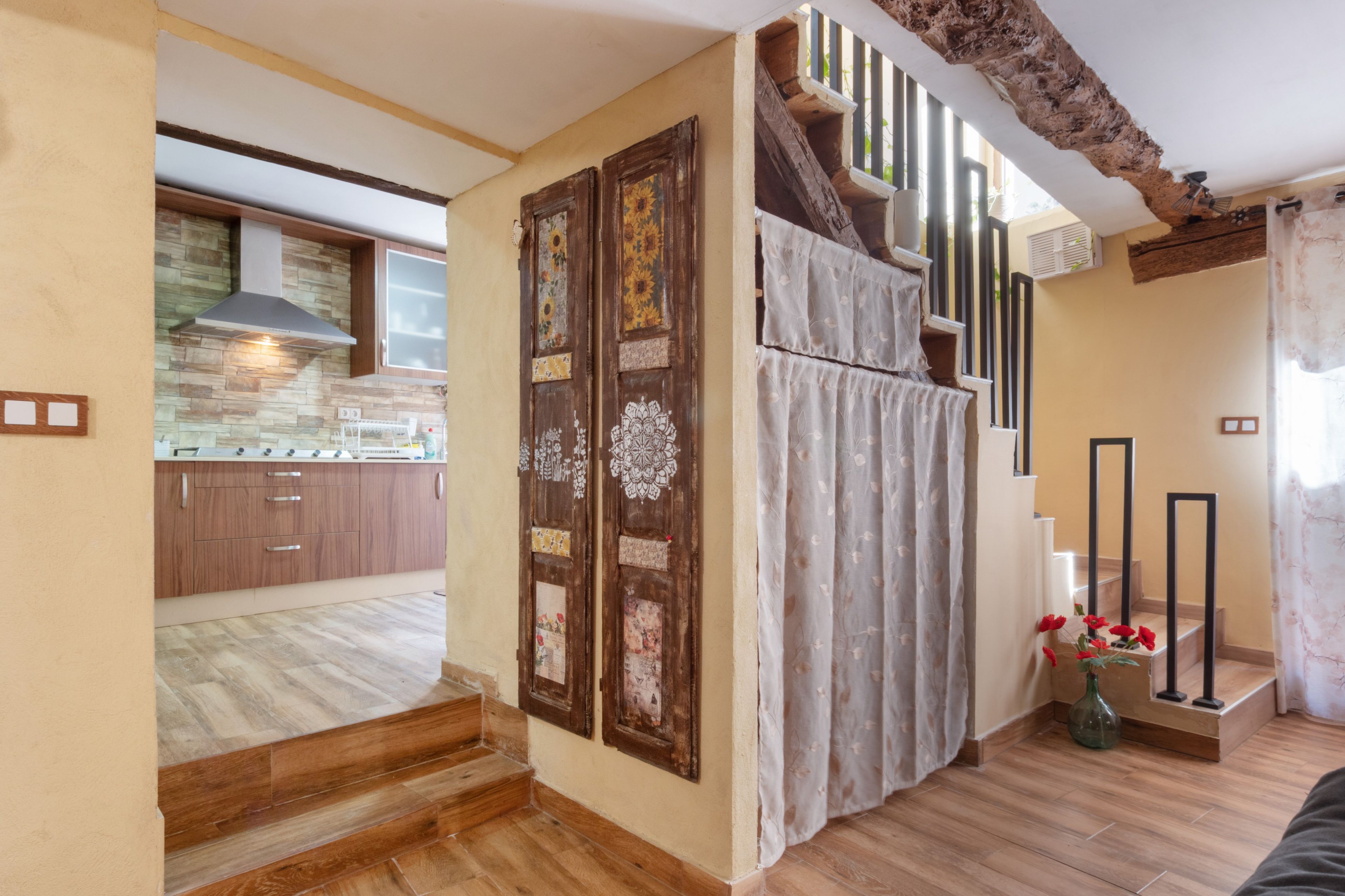 A kitchen with wooden cabinets and a stone backsplash, a staircase with a decorative door, and a view of the living area.