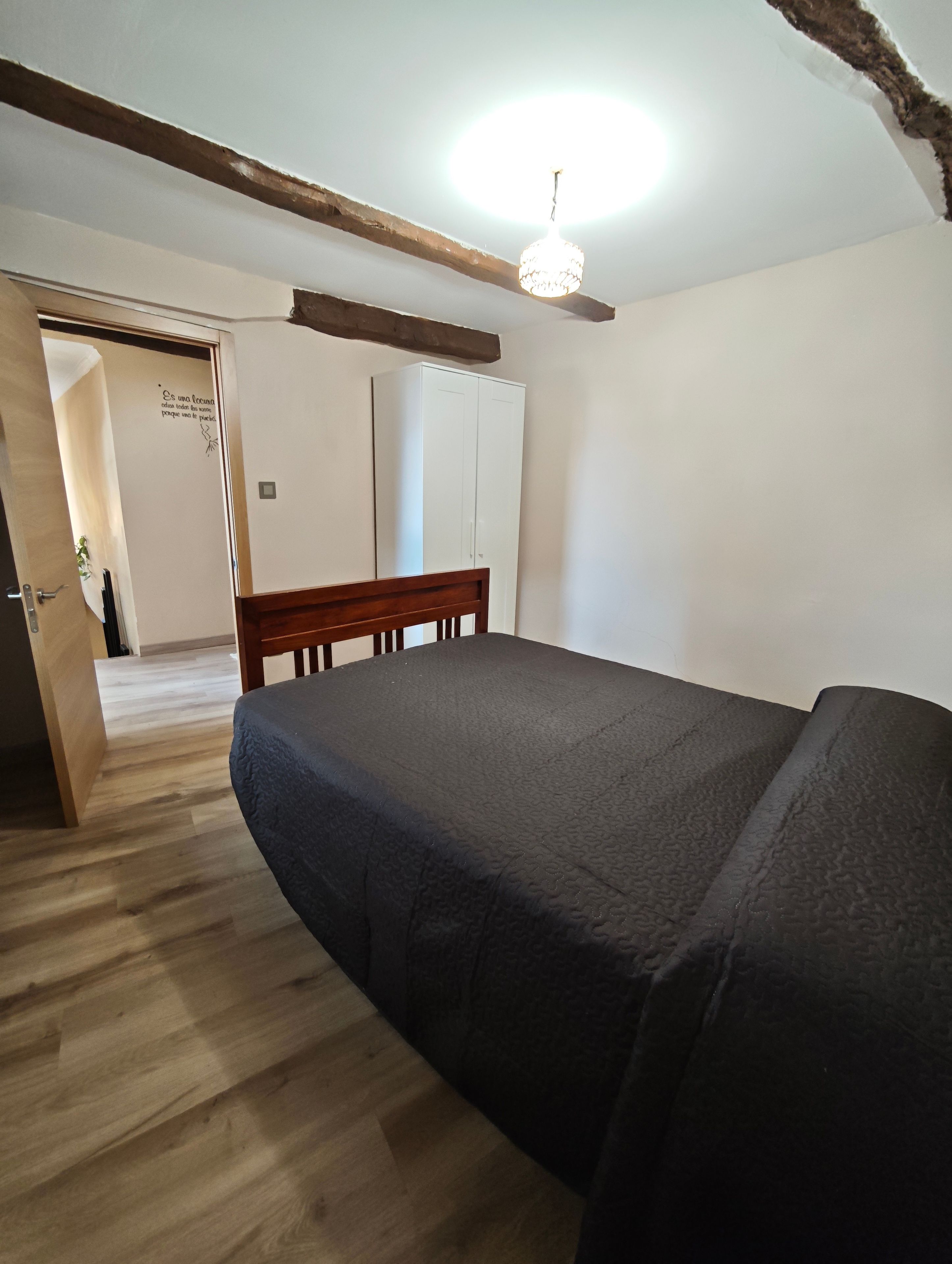 Un dormitorio con cama, piso de madera, armario y una puerta que da a otra habitación.