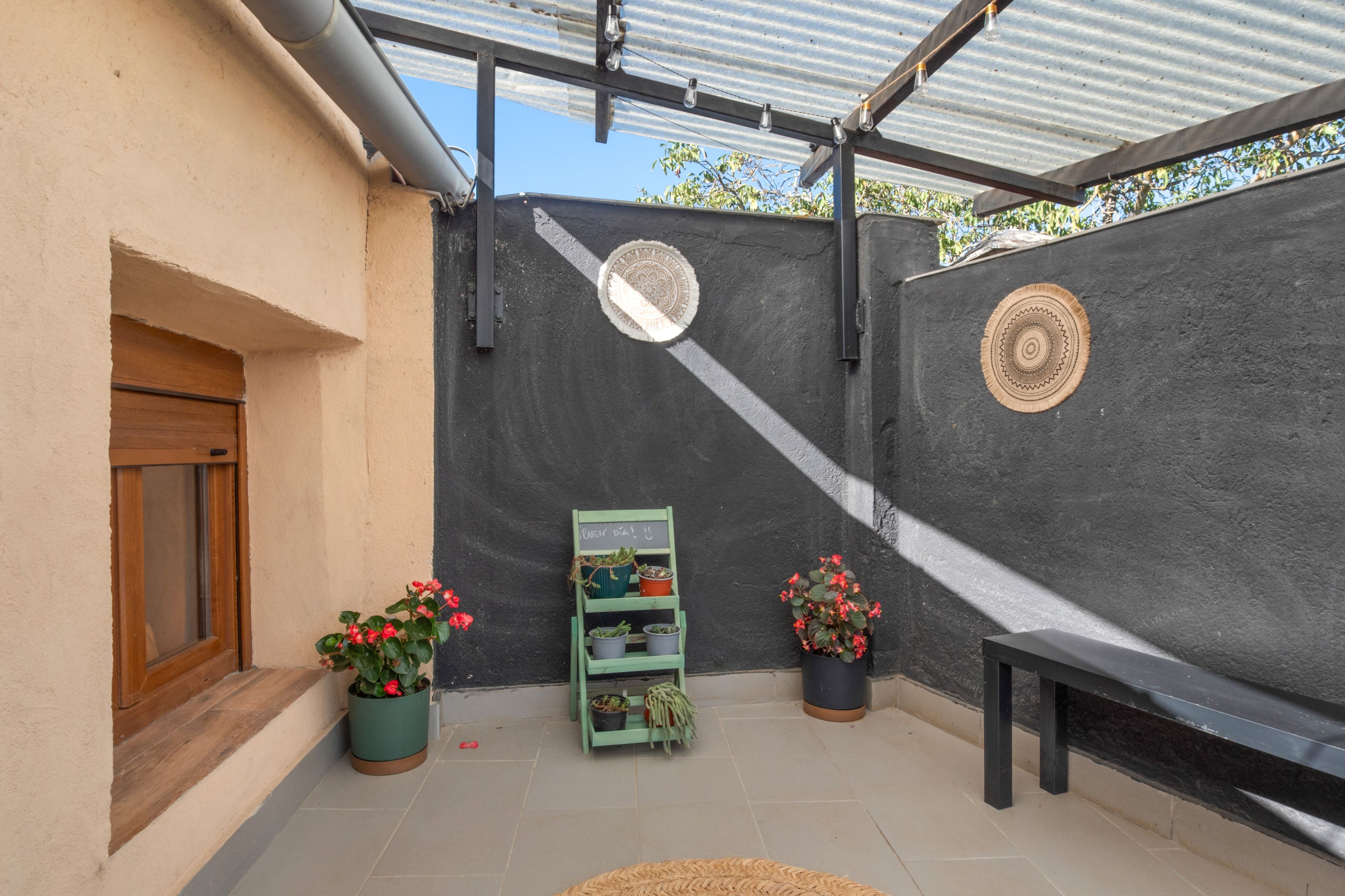 Un espacio exterior cubierto con un banco, plantas en macetas y un estante verde con herramientas de jardinería.
