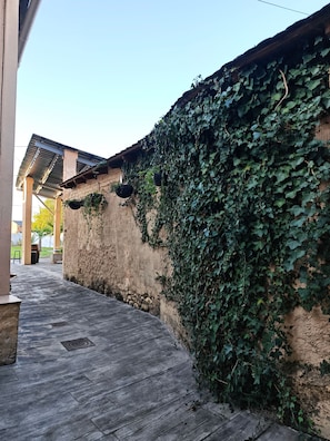 Un espacio exterior cubierto con suelo de madera, pared de piedra cubierta de hiedra y estructura de techo metálico.