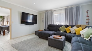 A modern living room with a sectional sofa, a flat-screen TV mounted on the wall, and a dining area with a table and chairs. - 58 Saltwater Crescent (Diamond Beach)