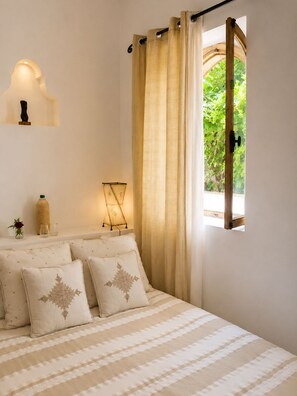 Une chambre à coucher avec un lit, des oreillers, une table de chevet avec une lampe, une fenêtre avec des rideaux et un luminaire fixé au mur.