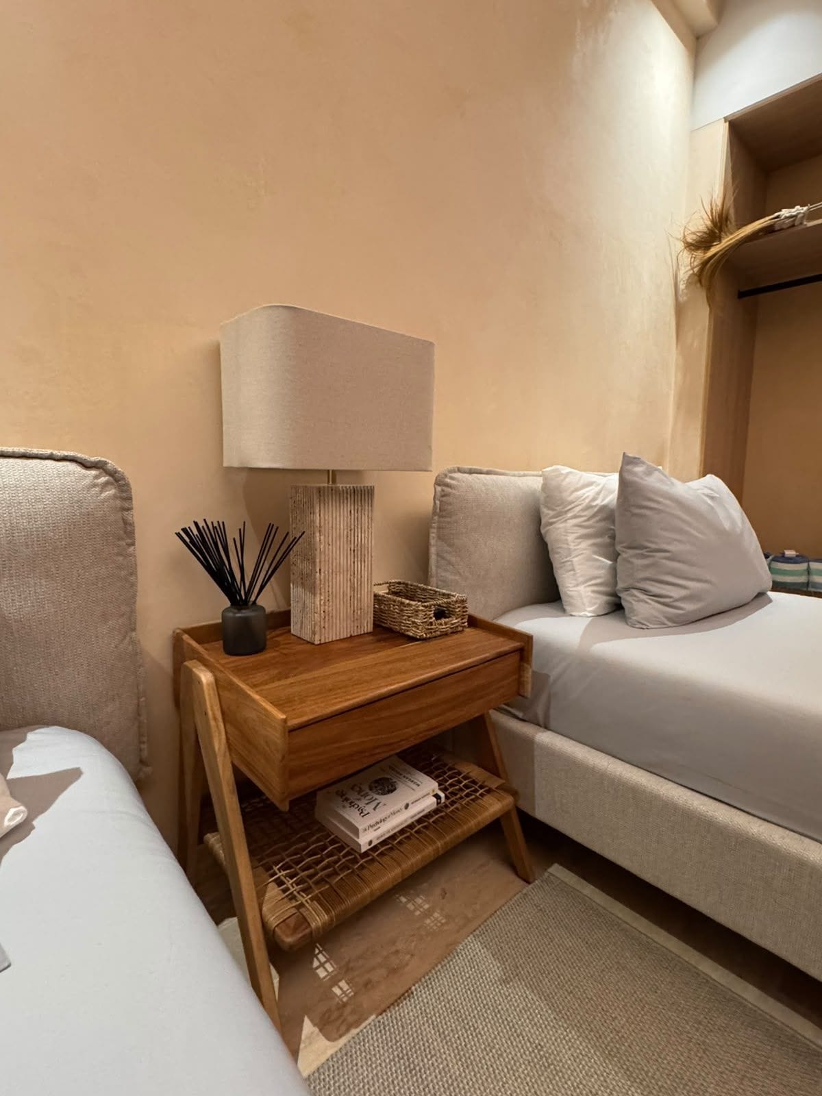 A neatly arranged bedroom with a bed, bedside table, lamp, and a small shelf with books and decorative items.