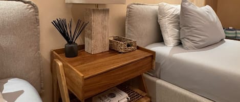 A neatly arranged bedroom with a bed, bedside table, lamp, and a small shelf with books and decorative items.