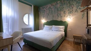 Un dormitorio con cabecera verde, ventana redonda, una silla, una mesita de noche y una pared con estampado floral.