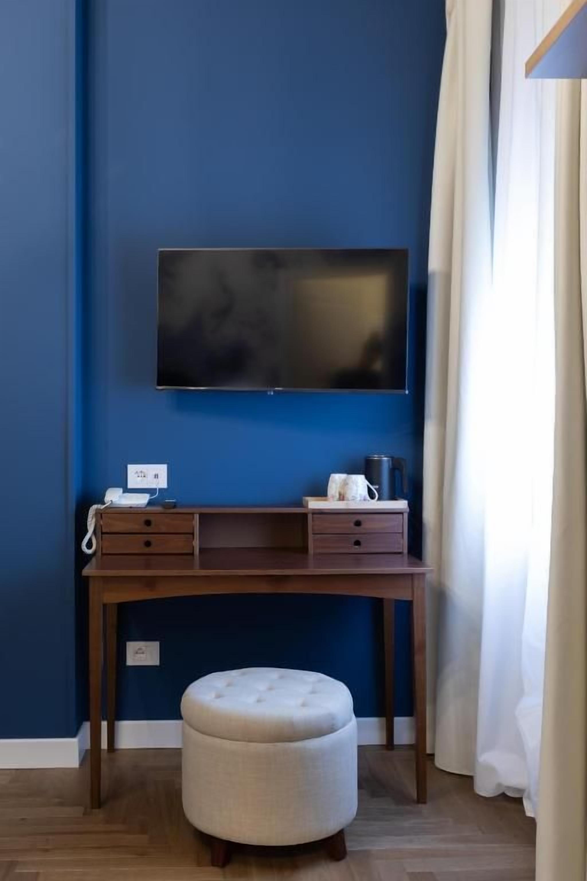 Una habitación con pared azul, un televisor de pantalla plana montado, un escritorio de madera con cajones, un otomano redondo pequeño y una cortina blanca.
