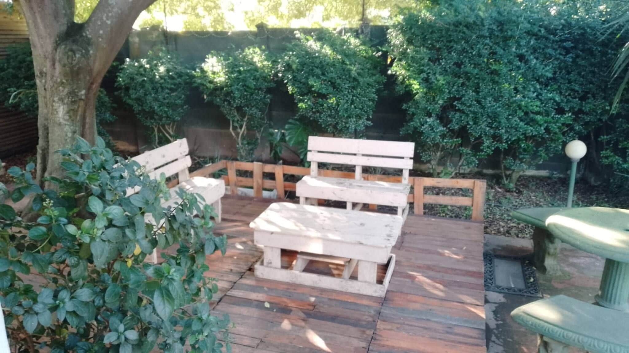 An outdoor seating area with a wooden bench and table, surrounded by greenery.