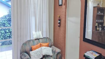 A room with a window, a fire extinguisher mounted on the wall, a chair with cushions, and a table with a stack of magazines.