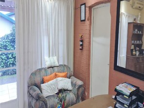 A room with a window, a fire extinguisher mounted on the wall, a chair with cushions, and a table with a stack of magazines. - Room-alexandra Bed & Breakfast Room 7 (Pietermaritzburg)