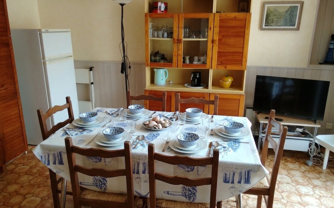 Ein Essbereich mit einem Tisch für vier Personen, umgeben von Holzsesseln. Ein Fernseher ist rechts an der Wand befestigt.