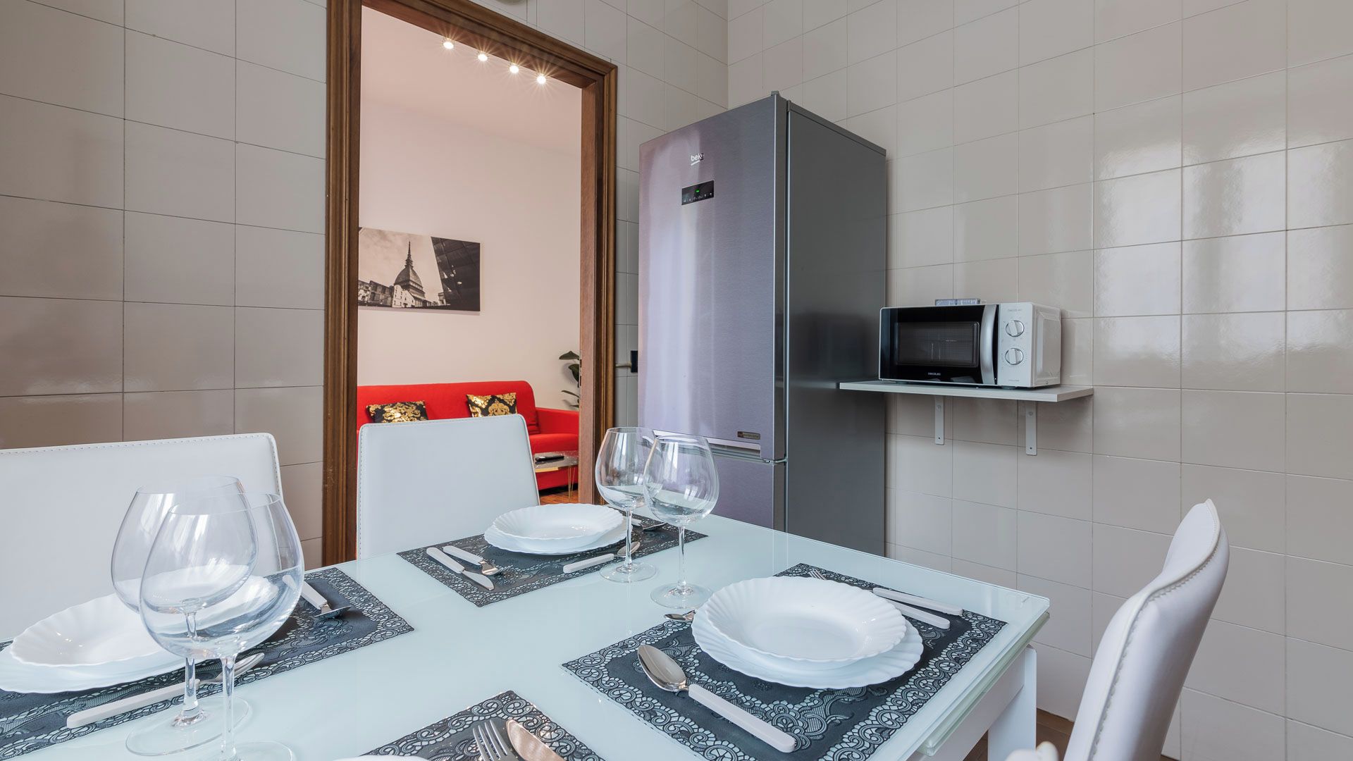A dining area with a table set for two, a microwave on a shelf, and a refrigerator in the background.