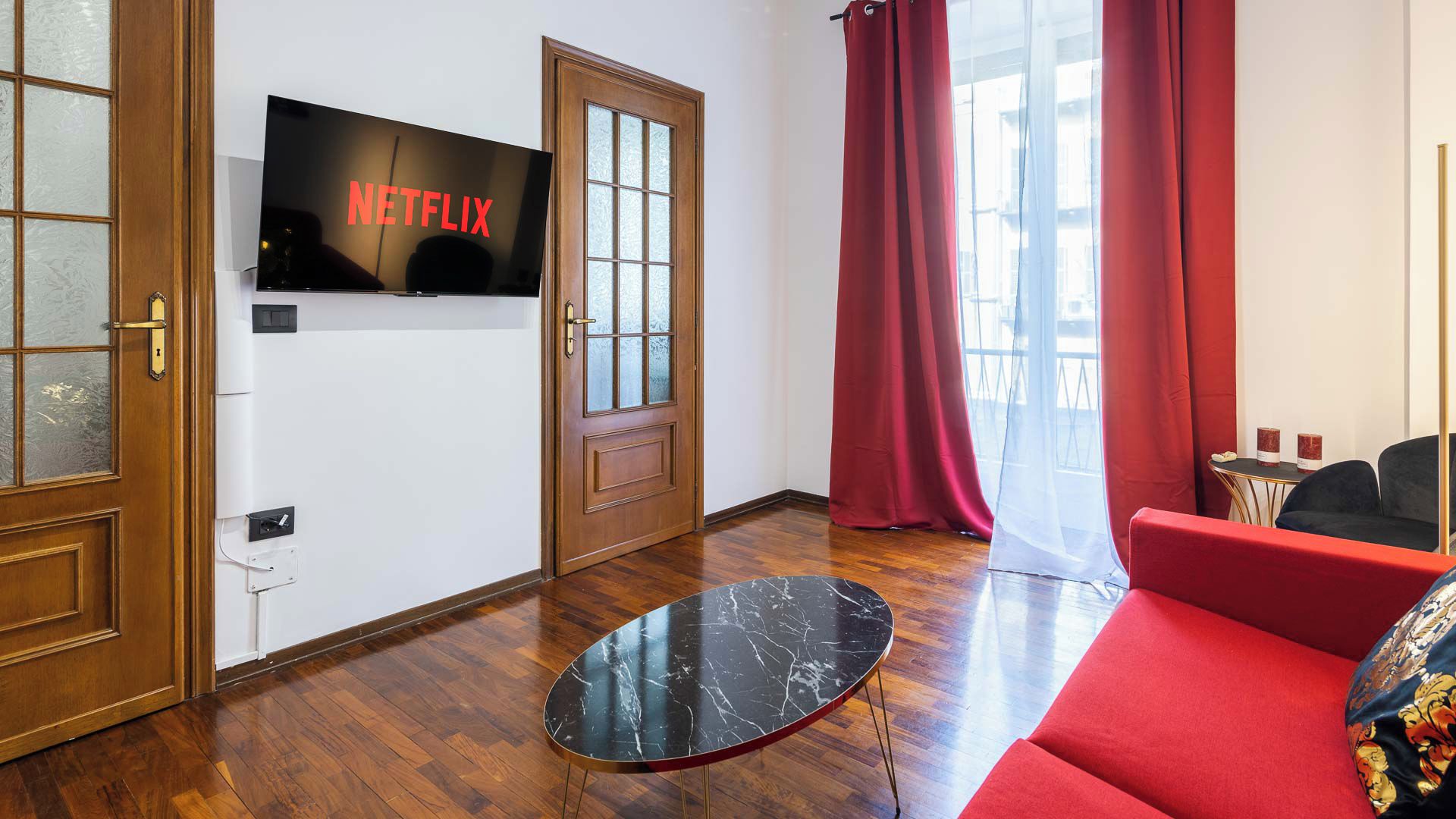 A modern living room with a flat-screen TV displaying the Netflix logo, a wooden door, a red sofa, and a marble coffee table.