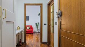 Un pasillo con una puerta de madera, un sofá rojo, una planta en maceta y un jarrón blanco sobre una mesa.