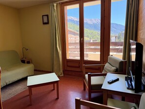 Een kamer met een bank, een stoel, een tafel, een televisie en uitzicht op bergen door een raam.