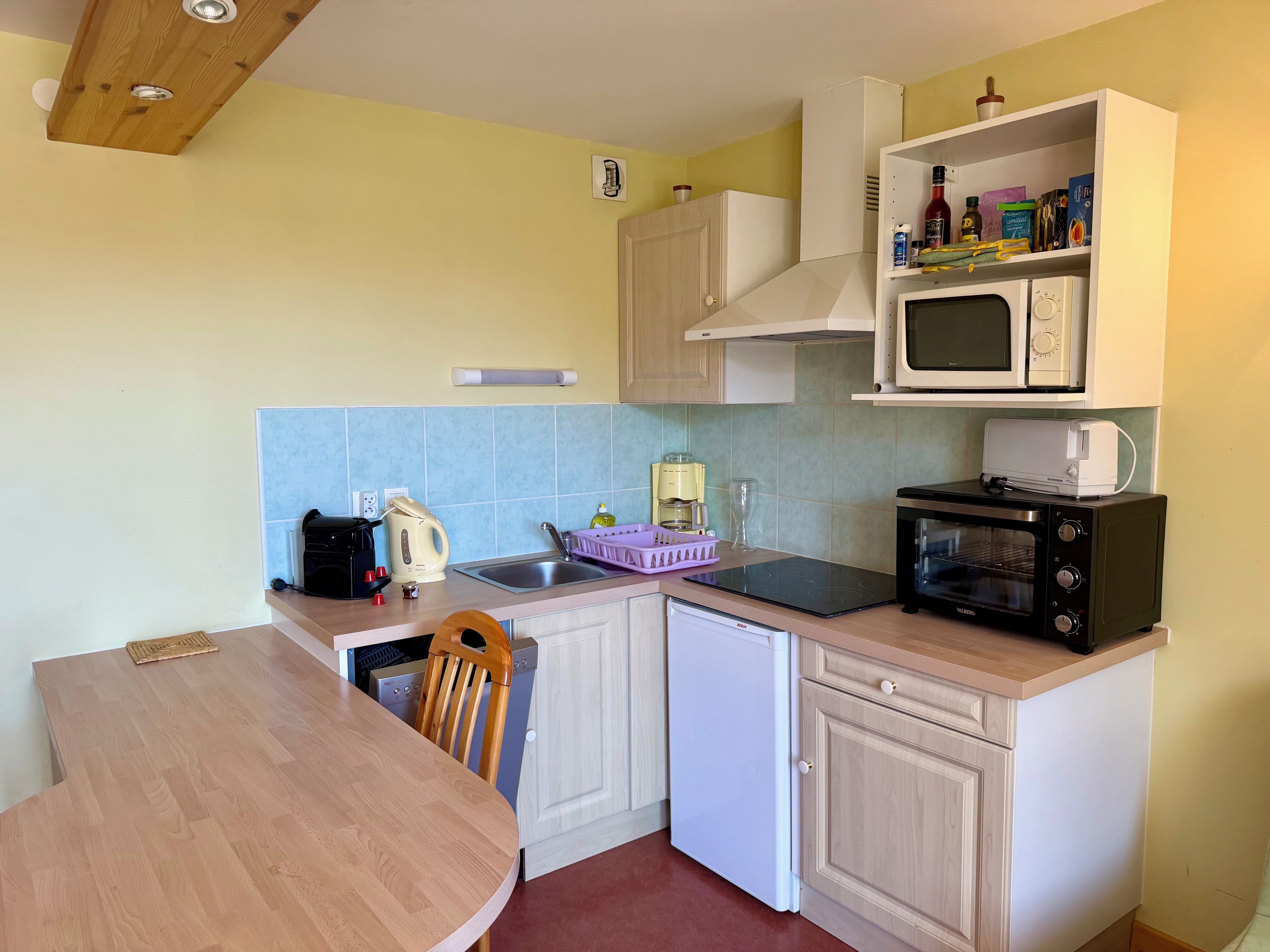 Een functionele keuken met een houten tafel, witte kasten, een magnetron, een broodrooster en een vaatwasser.