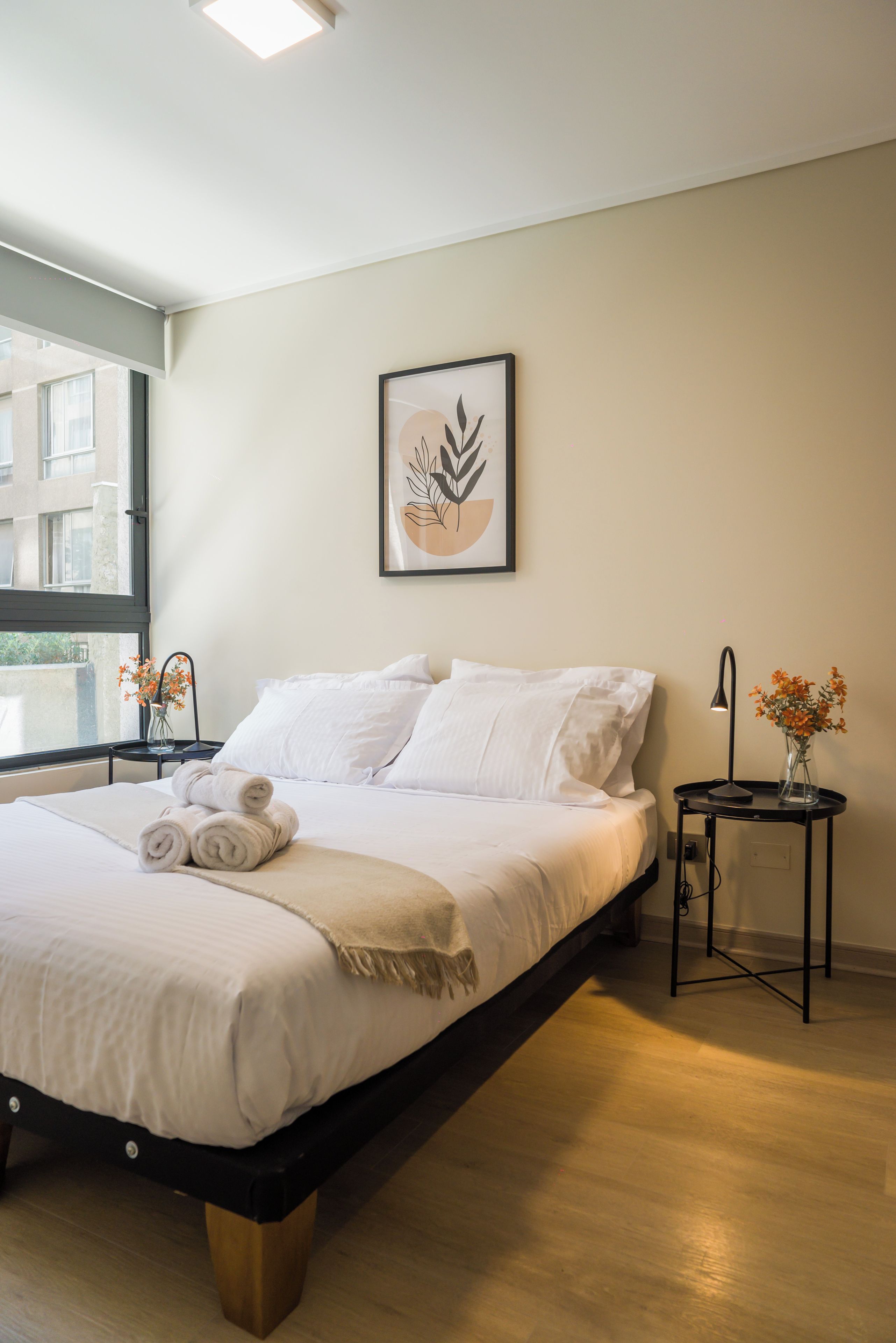 Une chambre à coucher moderne avec un lit, des oreillers, une table de chevet ornée d’un vase de fleurs et une œuvre d’art encadrée accroch ée au mur.