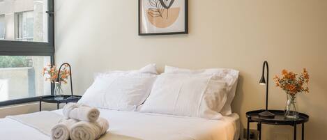 A modern bedroom with a bed, pillows, a bedside table with a vase of flowers, and a framed artwork on the wall.