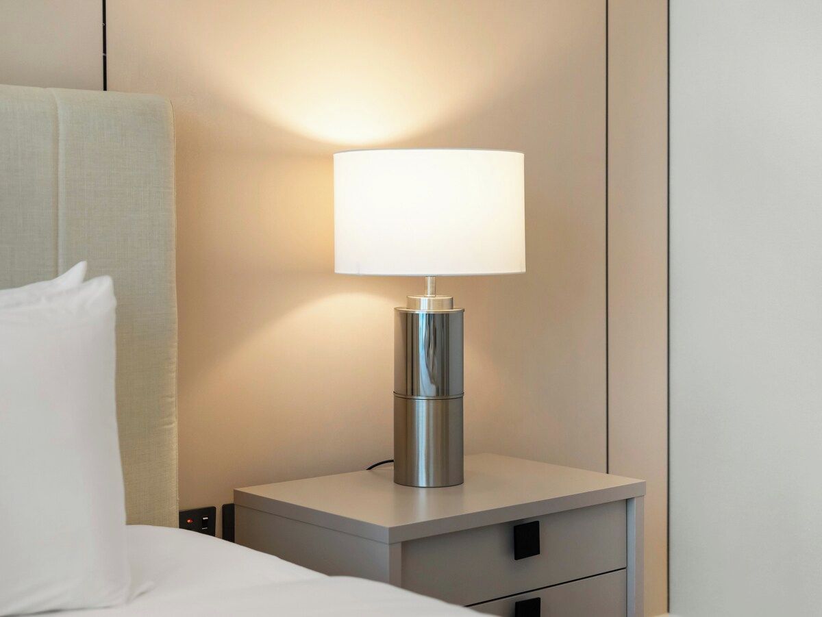 A modern bedside table with a lamp, a bed with white bedding, and a plain wall.