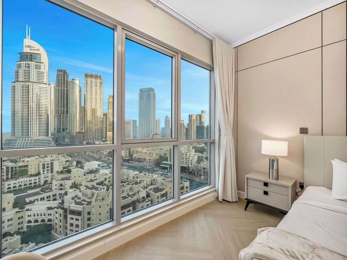 A hotel room with a large window, a bed, a nightstand, and a view of a cityscape.