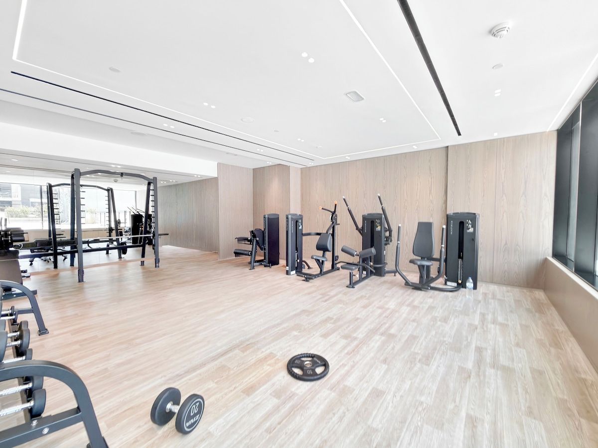Une salle de sport moderne équipée de divers appareils d’exercice, dotée d’un plancher en bois et de grandes fenêtres.
