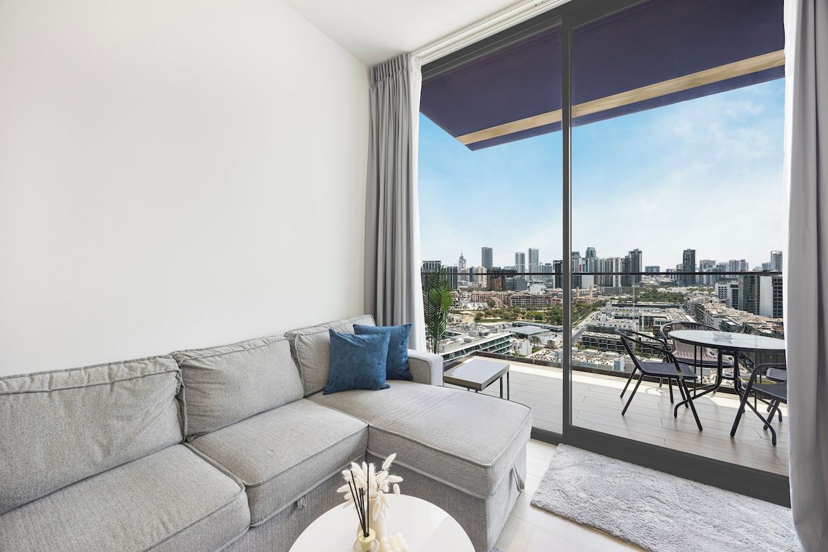 A modern living room with a grey sofa, a small white table, and a city view through large windows.