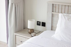 A neatly made bed with white bedding, a bedside lamp, a small decorative item, and a power outlet on a wooden nightstand. - City Views / Pool & Gym / Near Circle Mall JVC (Dubai)