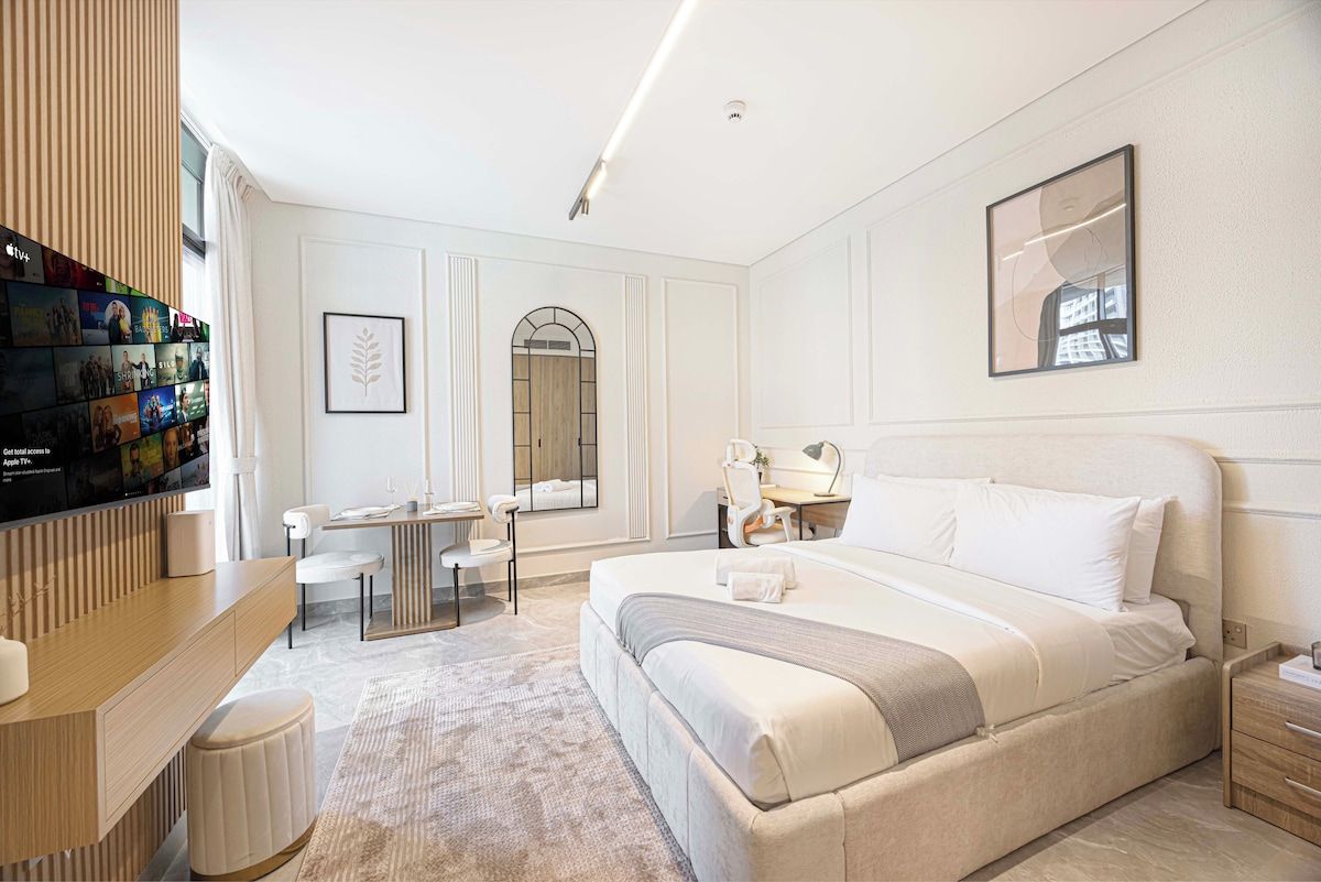 Une chambre d’hôtel avec un grand lit, un bureau avec une chaise, une télévision et une étagère murale remplie de livres.