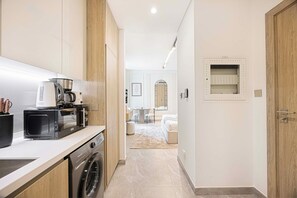 Une cuisine moderne équipée d’une machine à café, d’un four à micro-ondes et d’un lave-linge. Un couloir mène à une chambre avec un lit et une petite table.