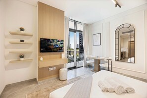 Ein Hotelzimmer mit einem Bett, einem Fernseher, einem kleinen Tisch, einem Stuhl, einem Spiegel und einem Balkon mit Blick auf Gebäude.