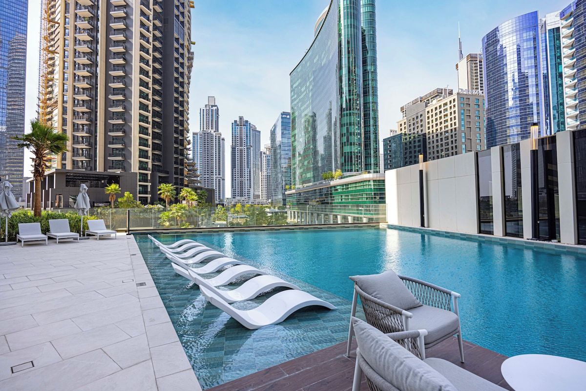 A rooftop pool with lounge chairs and a modern building complex in the background.