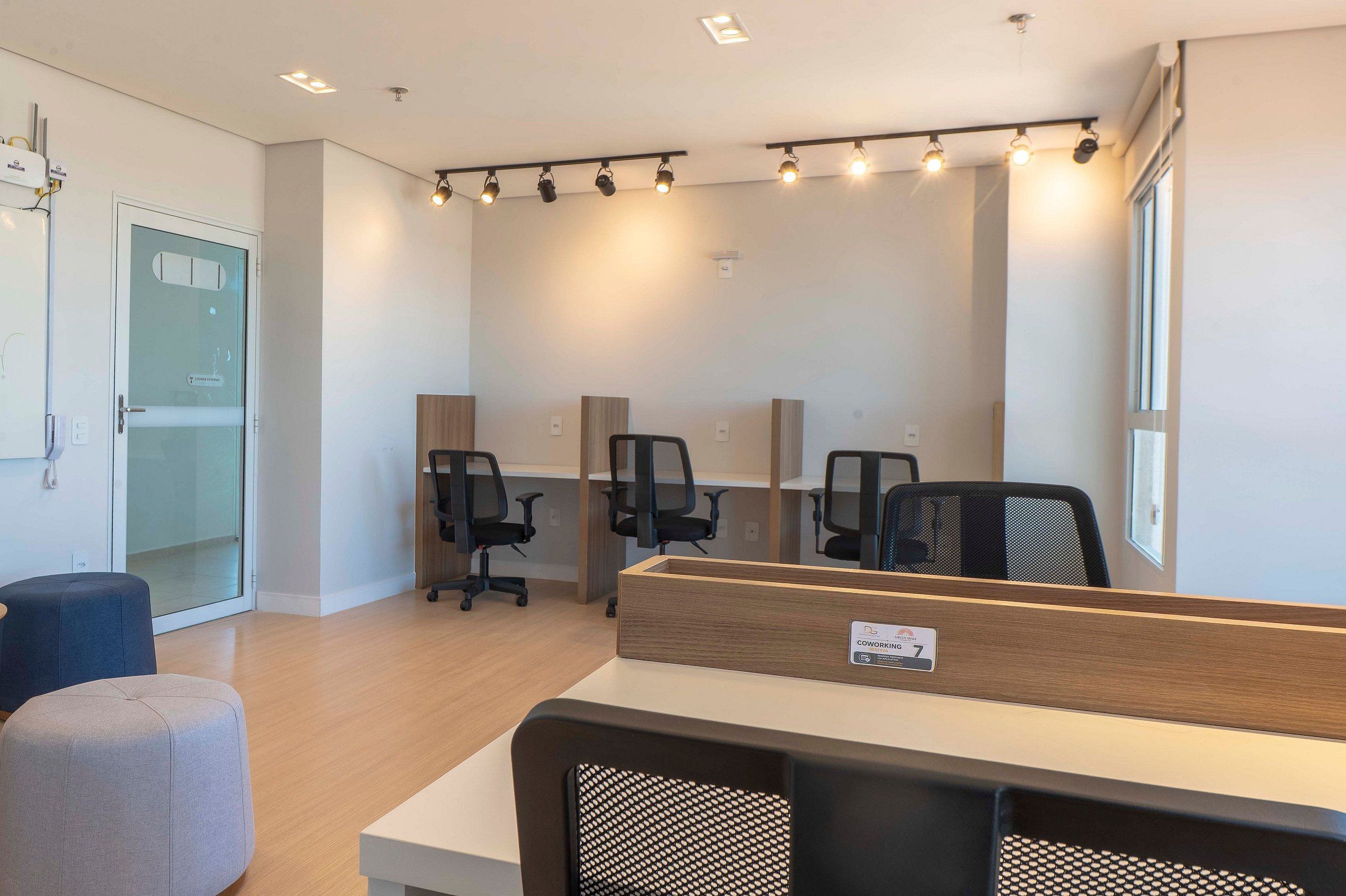 A modern office space with cubicles, desks, and chairs. There are ceiling lights and a glass door on the left.