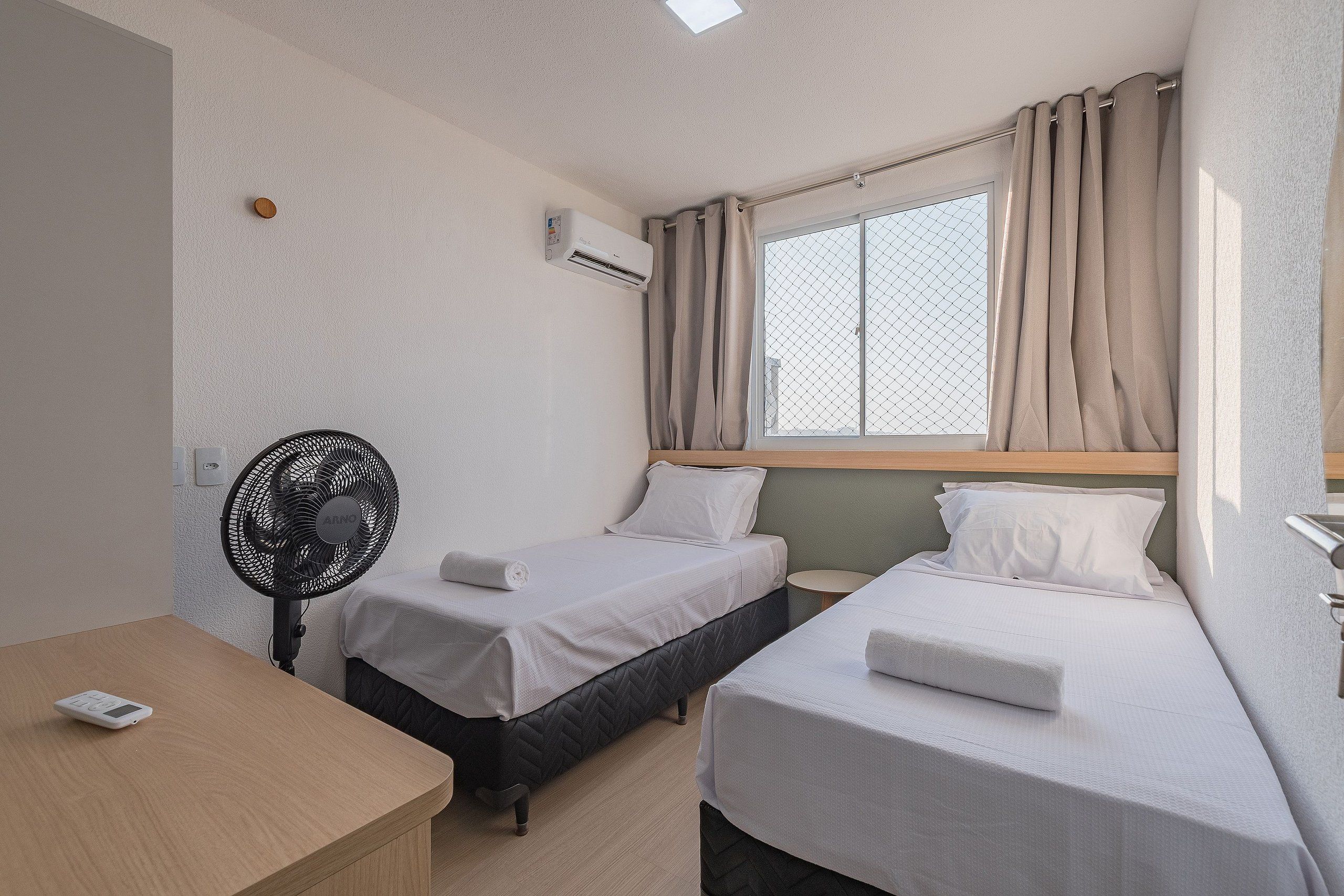 Habitación de hotel con dos camas individuales, una mesita de noche, un ventilador, una ventana con cortinas e instalación de aire acondicionado.