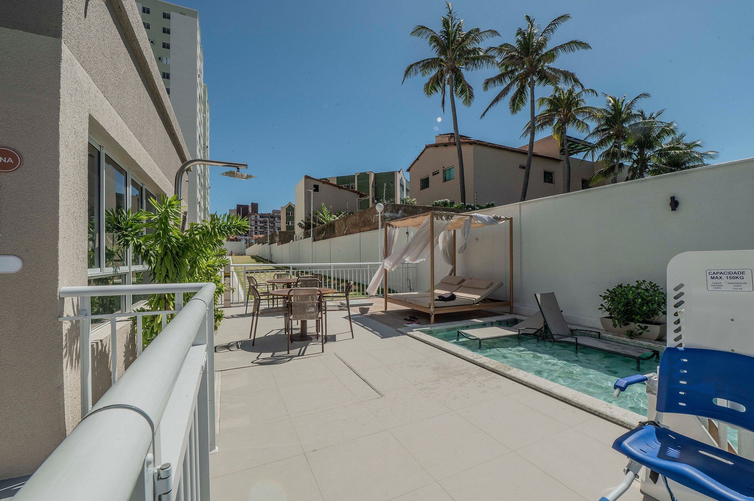 A pool area with lounge chairs, a covered seating area, and a view of palm trees and buildings.