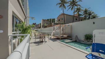 Zona de piscina con sillas de descanso, un área de estar cubierta y vistas a palmeras y edificios.