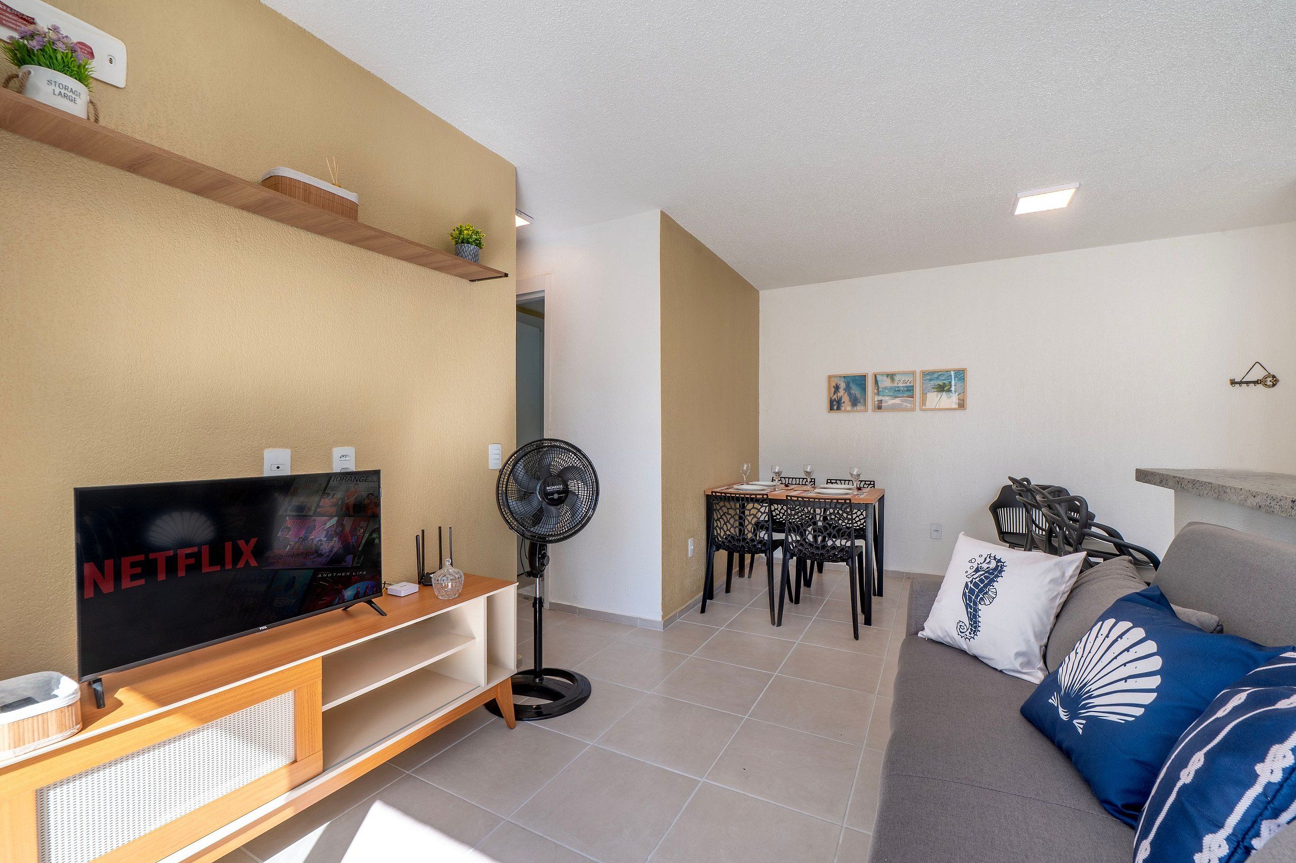A living room with a TV, a fan, a dining area, and a sofa with decorative pillows.