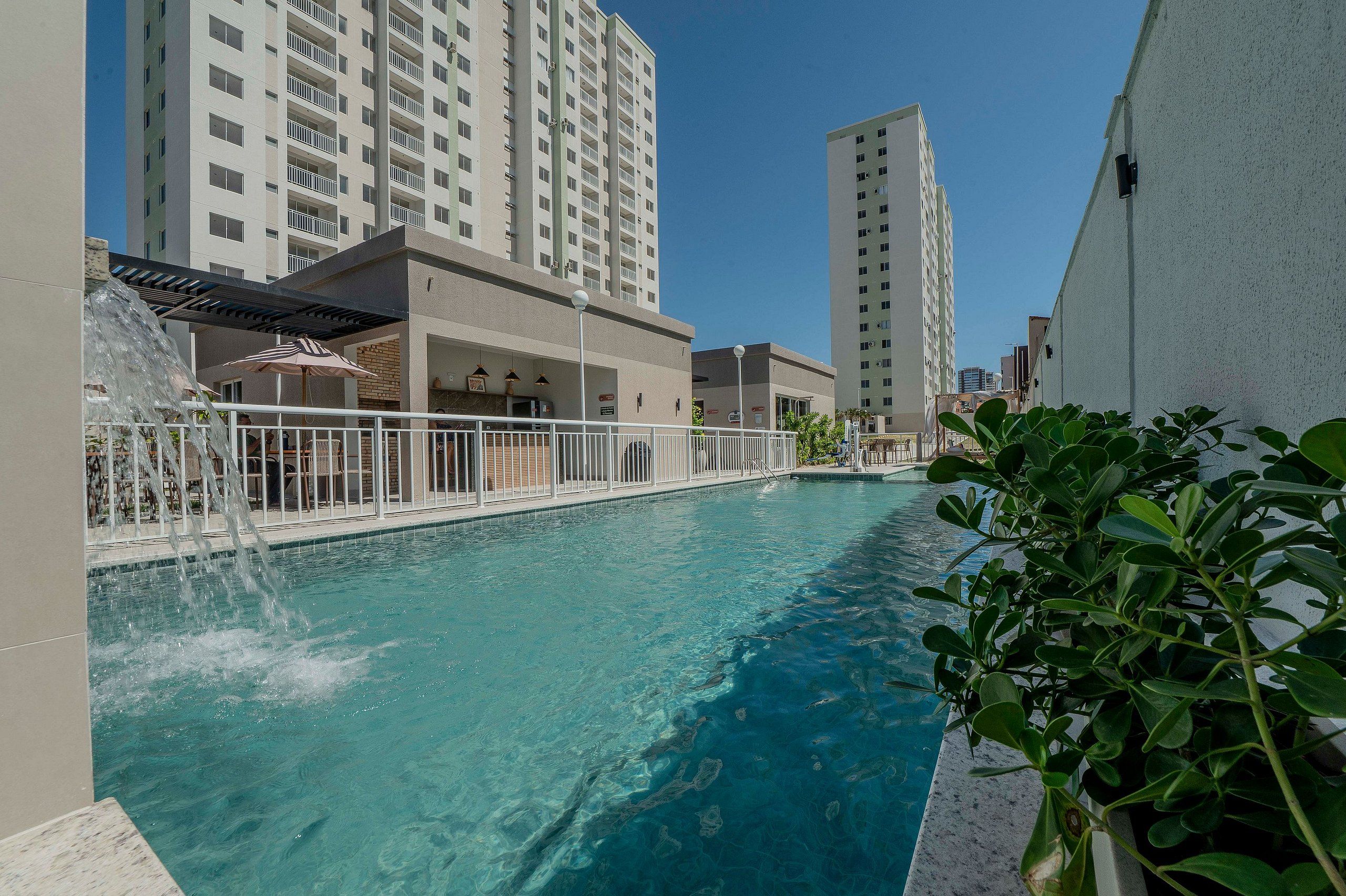 A swimming pool with a water fountain, surrounded by a white fence, with tall buildings in the background.