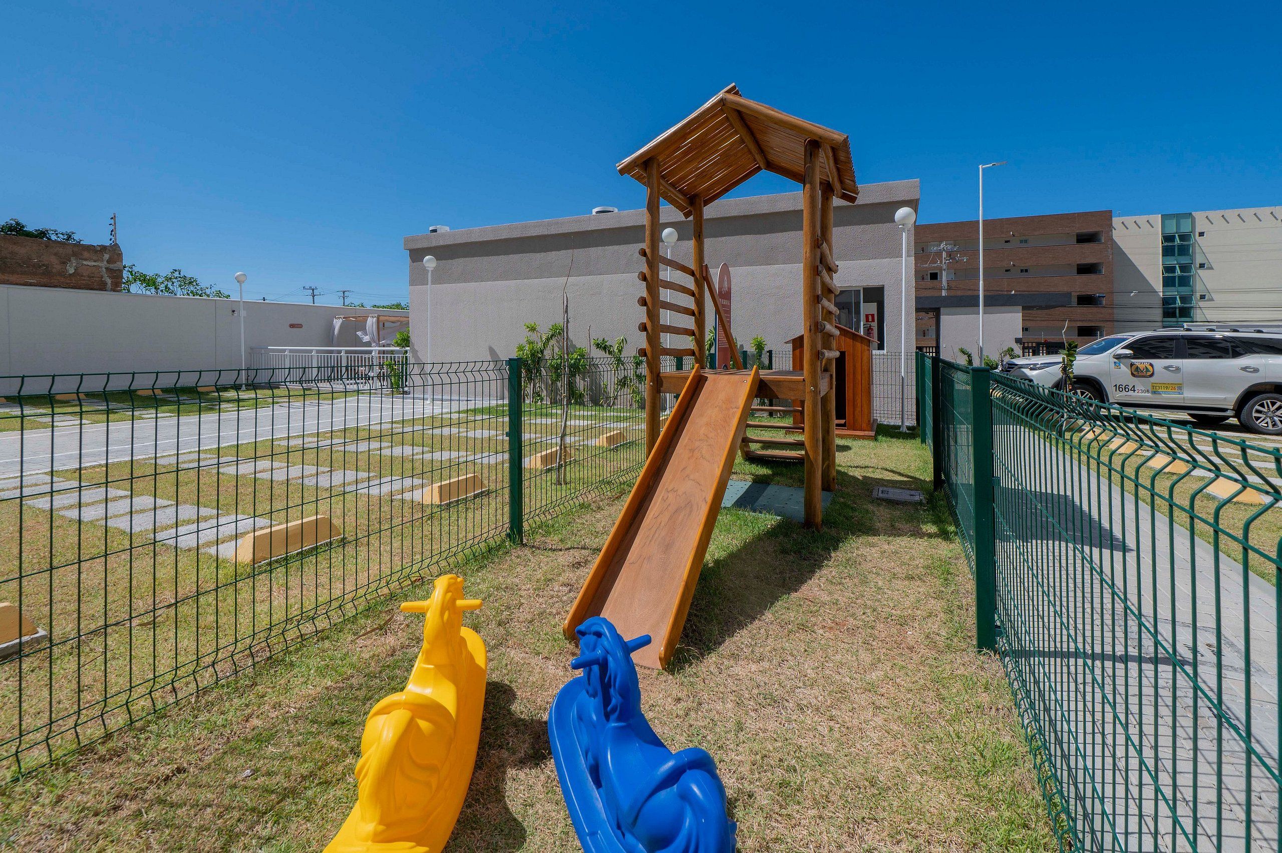 Uma área de recreação infantil com escorrega e estrutura de escalada, cercada por uma grade e com um edifício ao fundo.