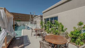 Zona junto a la piscina con mesa y sillas de madera, un espacio de descanso techado y vista a un edificio con piscina.