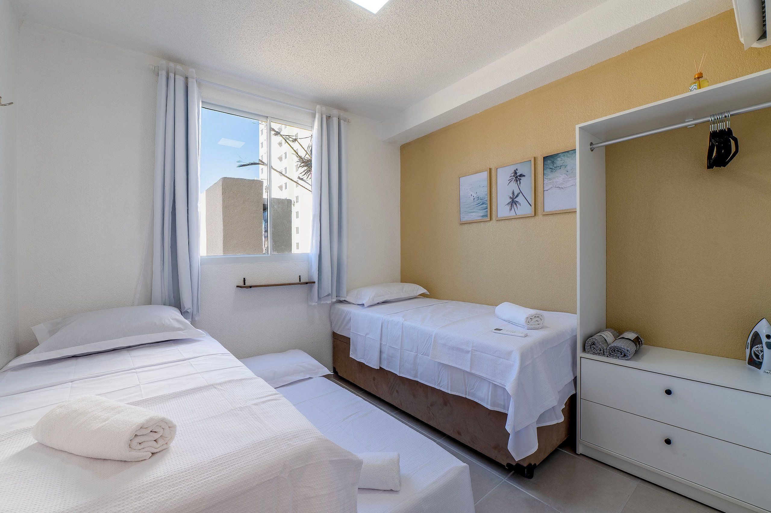 Habitación de hotel con dos camas, una ventana con cortinas, una pared con cuadros enmarcados y un aparador blanco con un planchador.