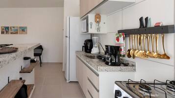 Una cocina compacta con estufa a gas, microondas y cafetera. Hay utensilios colgando y un pequeño estante con una botella.