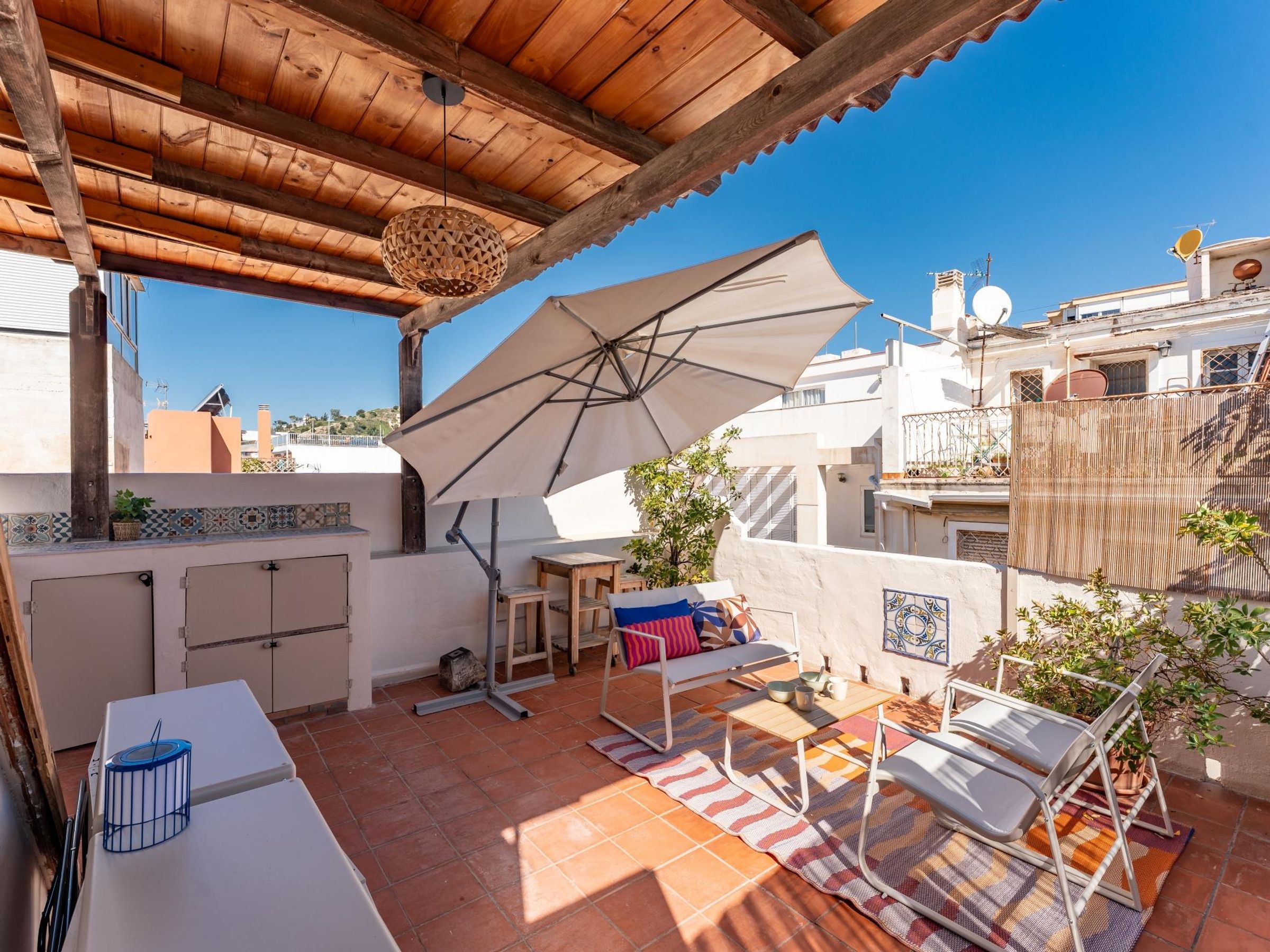 A rooftop patio with a wooden ceiling, a large umbrella, and a seating area with cushions.