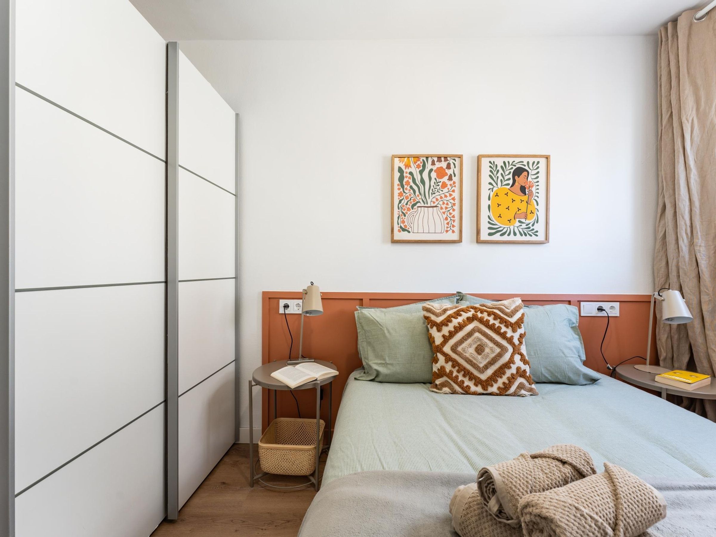 Een slaapkamer met een bed, kussens, een nachtkastje met een lamp, een wandplank en twee ingelijste kunstwerken.